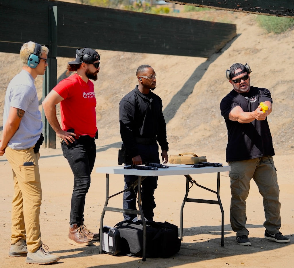 4 men standing outside at a gun range. One man is holding a dummy gun demonstrating how to grip the gun. The other three men are watching the demonstration. All the men are wearing pants, a tshirt, ear and eye protection. it is a sunny day. 